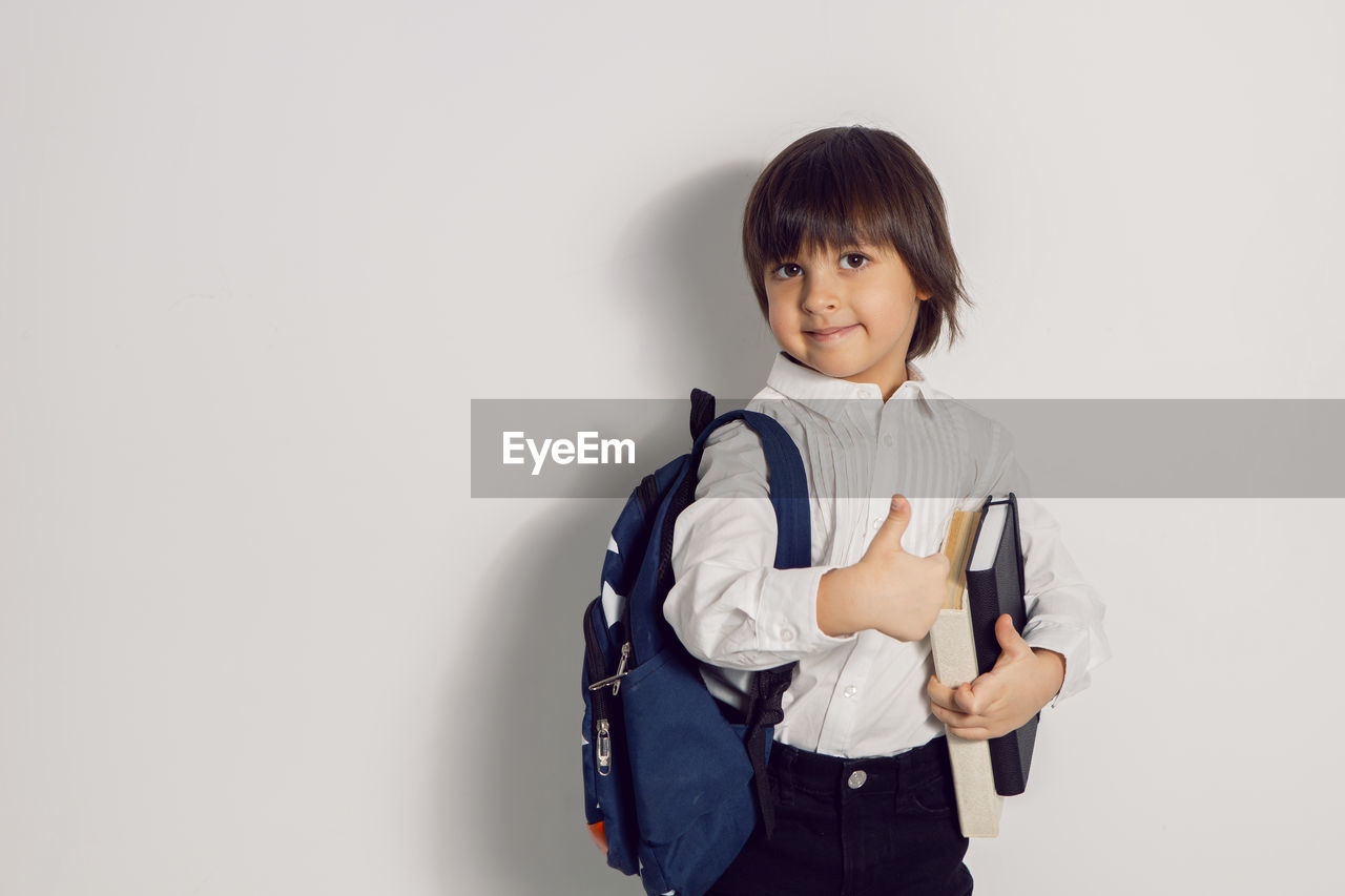 Child boy with a book textbook and backpack stands on a white background thumbs up