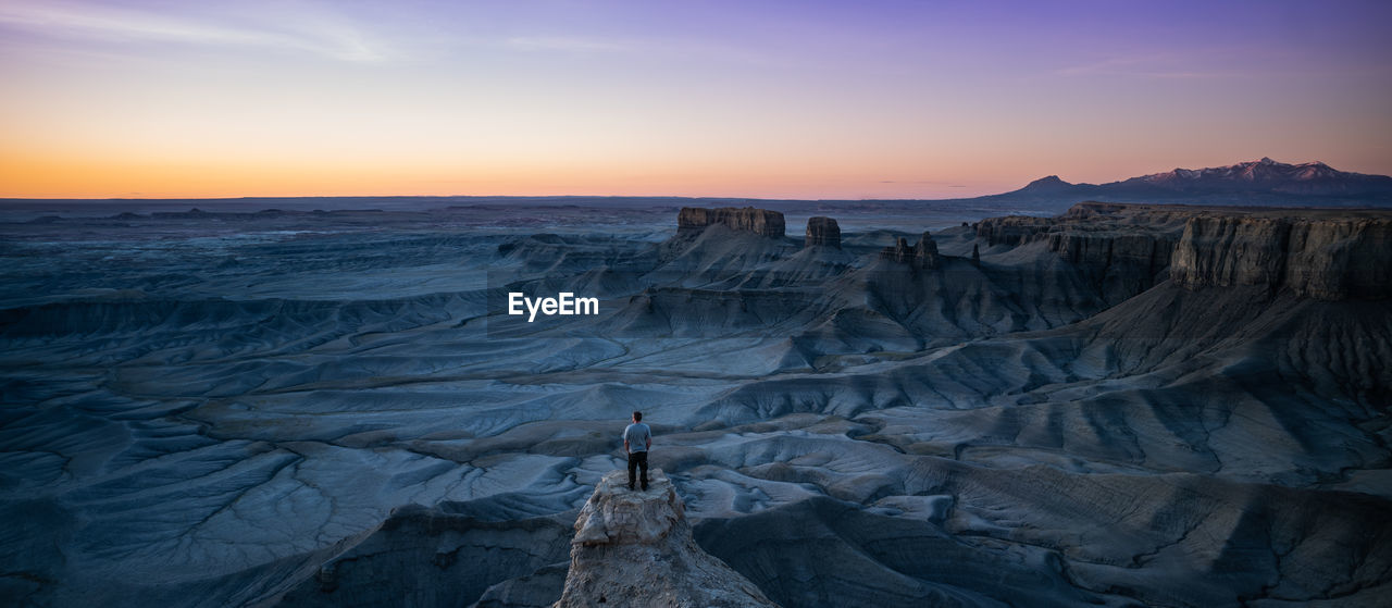 Rear view of man standing on rock formations against sky during sunset