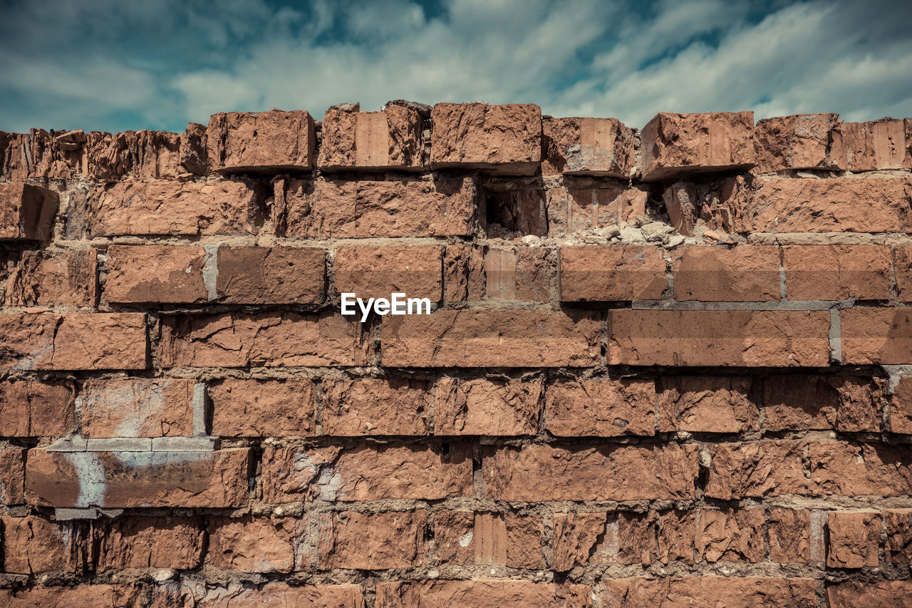 Low angle view of brick wall against cloudy sky during sunny day