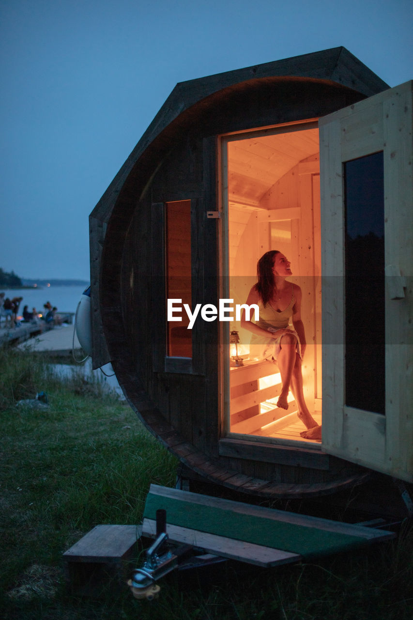 Full length of smiling woman sitting in sauna cabin by sea against sky