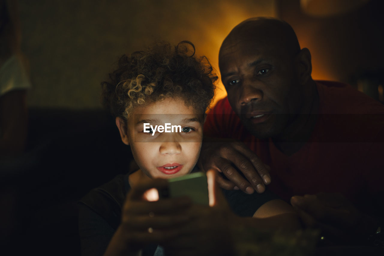 Boy using smart phone by father in living room at night