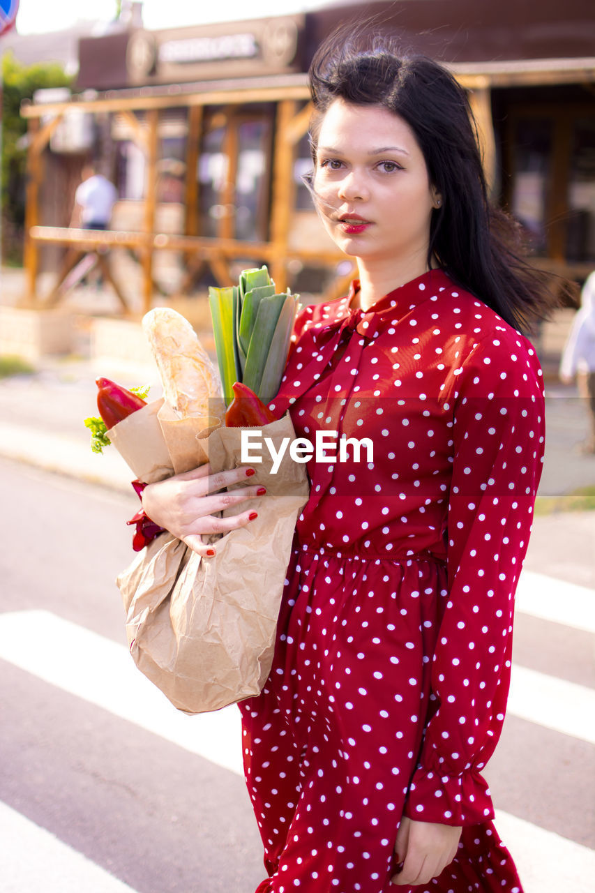 women, one person, adult, young adult, city, polka dot, smiling, street, happiness, lifestyles, clothing, architecture, city life, female, fashion, emotion, red, pattern, flower, standing, portrait, cheerful, pink, holding, shopping, outdoors, nature, bag, hairstyle, city street, retail, casual clothing, looking, three quarter length, shopping bag, consumerism, positive emotion, looking at camera, dress, food, flowering plant, food and drink, brown hair, day, long hair, spotted, leisure activity