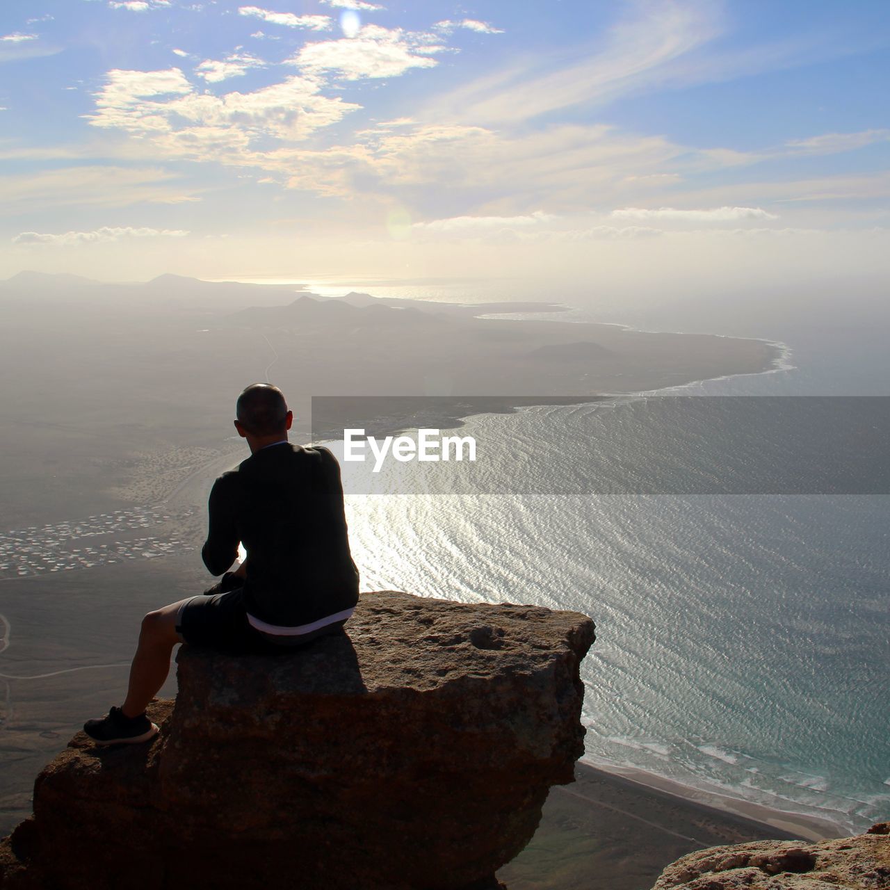 Rear view of man looking at sea while sitting on rock against sky