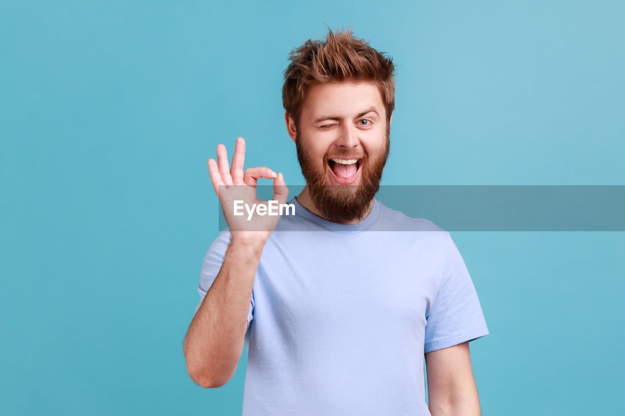 blue, smiling, happiness, colored background, studio shot, one person, finger, emotion, portrait, adult, men, young adult, casual clothing, beard, facial hair, blue background, cheerful, positive emotion, indoors, person, teeth, smile, copy space, hand, gesturing, waist up, looking at camera, t-shirt, front view, fun, standing, human face, redhead, brown hair, sign language, laughing, excitement, joy, clothing, teenager, looking, thumbs up, communication, humor, lifestyles