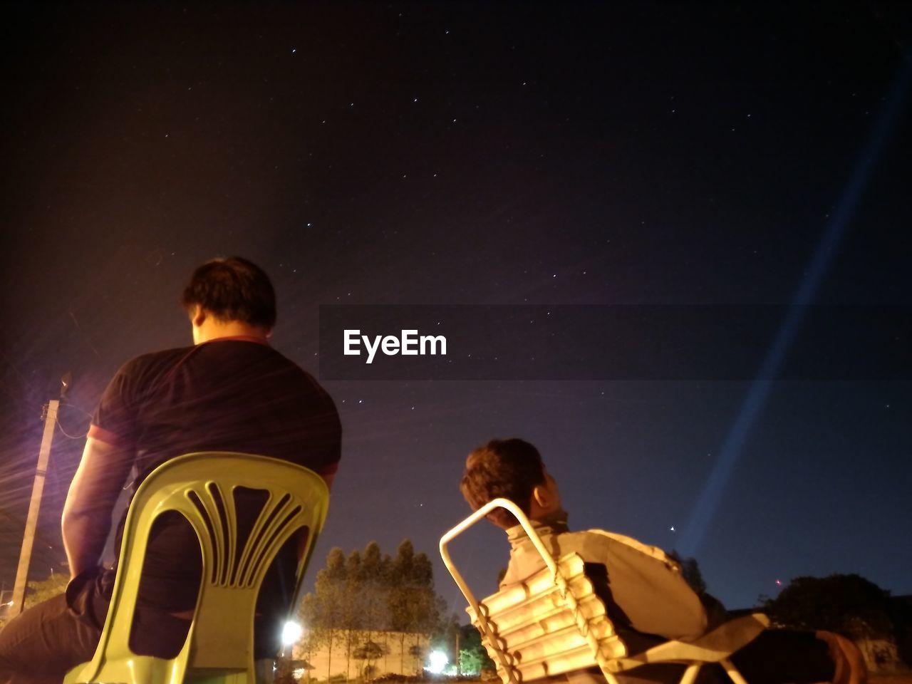 REAR VIEW OF MAN STANDING BY ILLUMINATED LIGHT AGAINST SKY