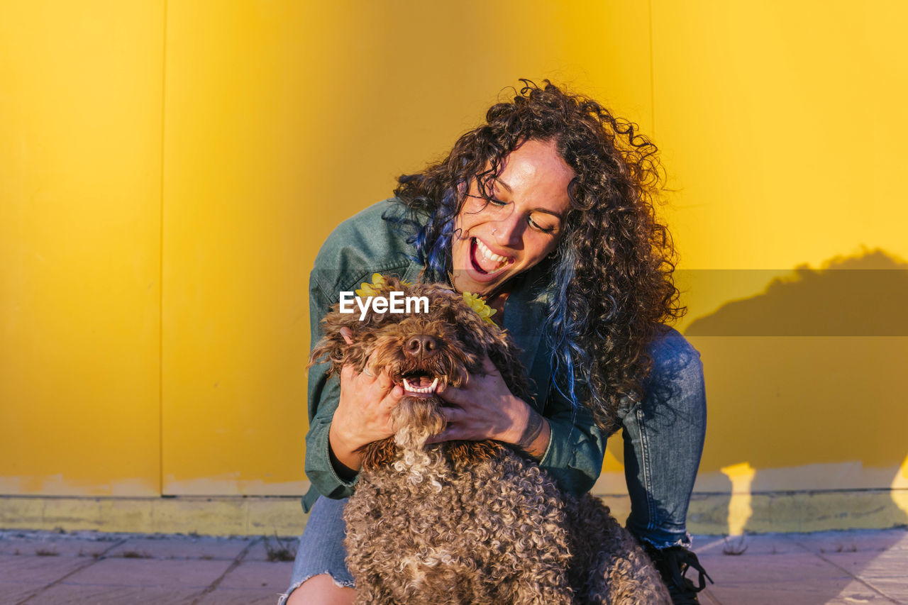 Happy woman playing with water dog in front of yellow wall