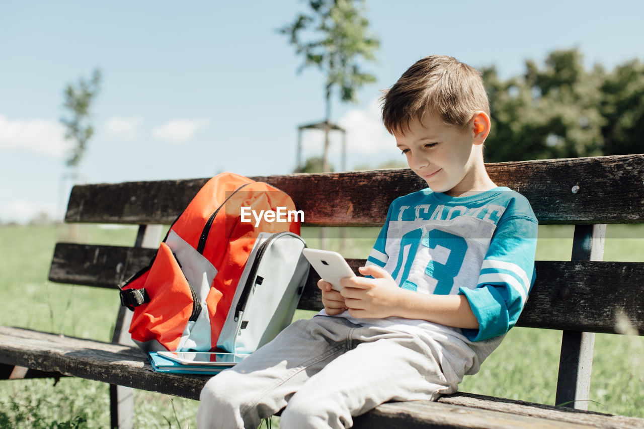 Boy using mobile phone while sitting on bench in park during sunny day