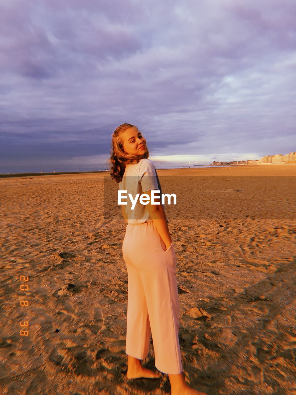 Full length rear view portrait of young woman standing on beach against cloudy sky