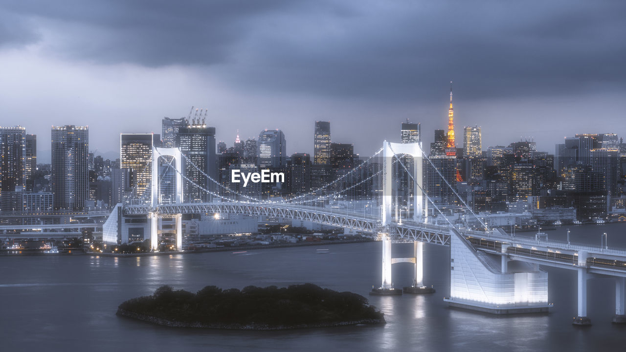 Illuminated bridge over river by buildings against sky