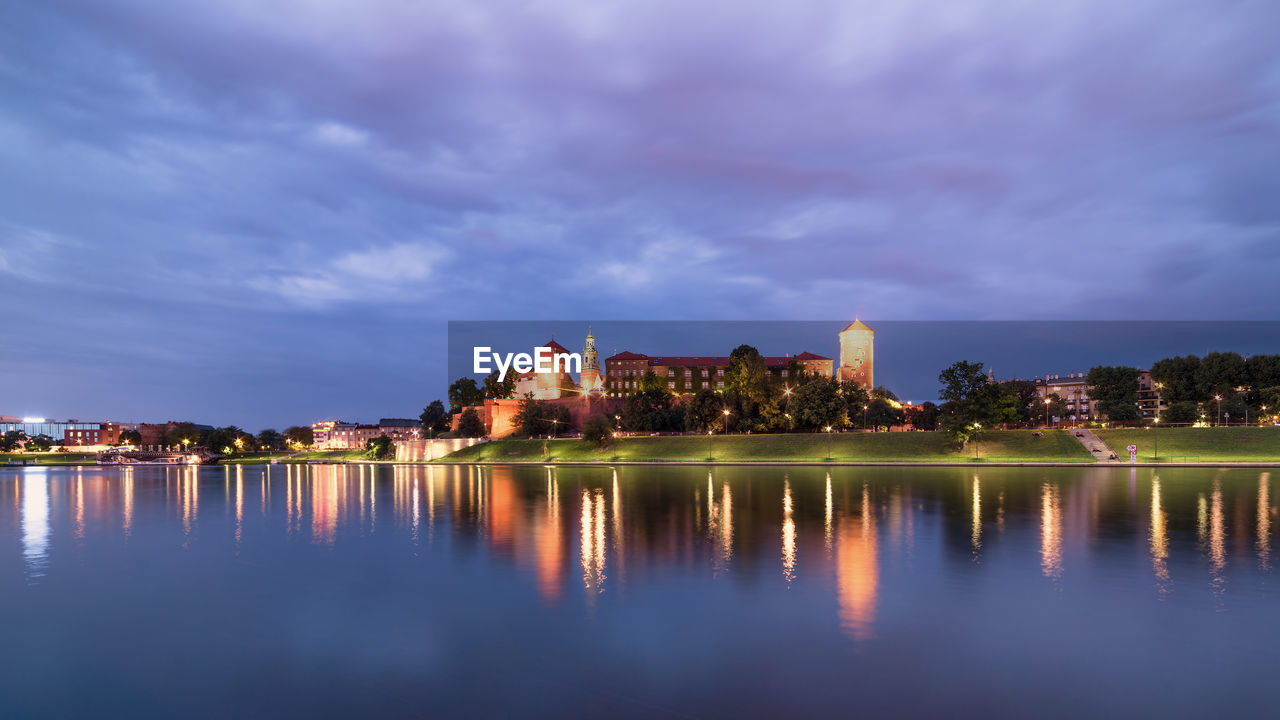 Illuminated buildings by river against cloudy sky