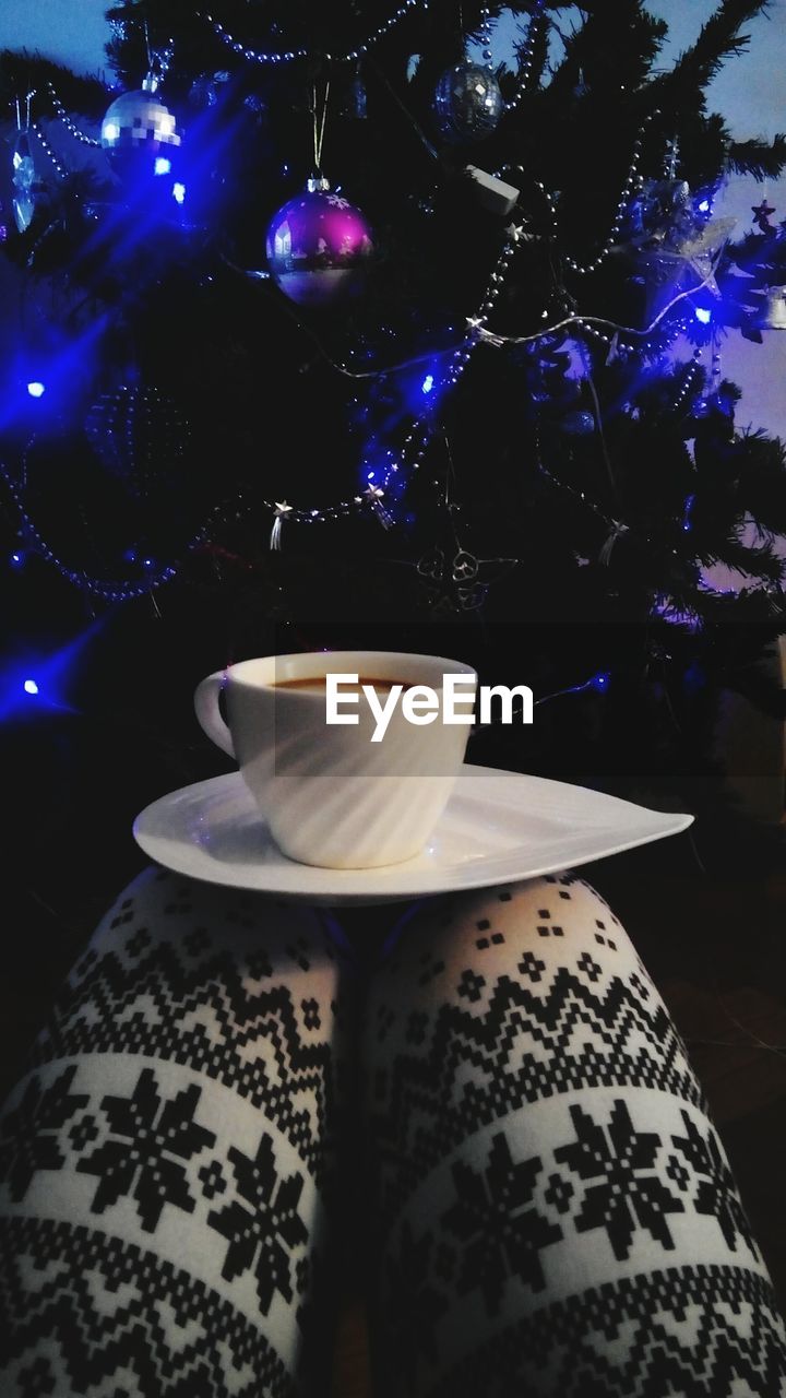 Low section of woman with coffee cup against christmas tree at home