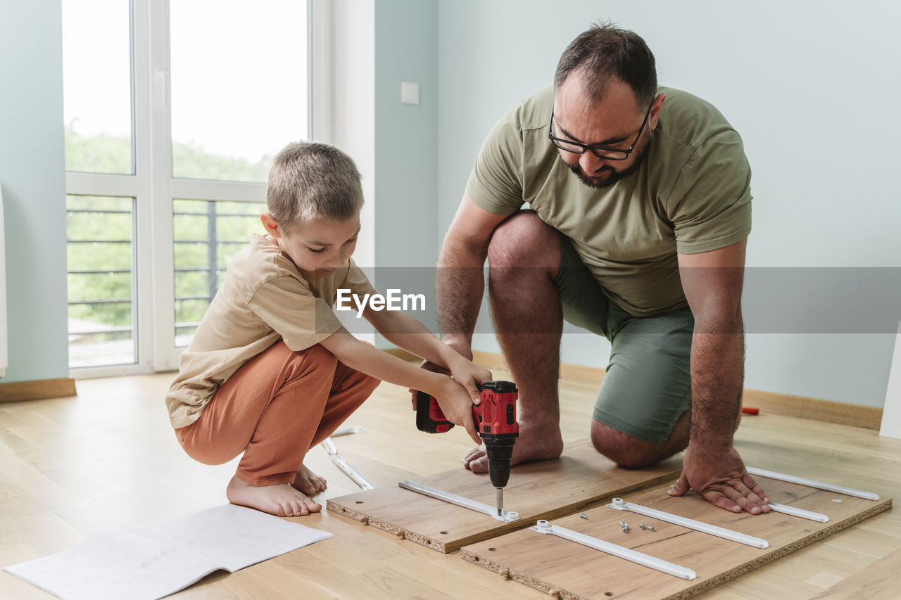 Father with son using drill on plank at home