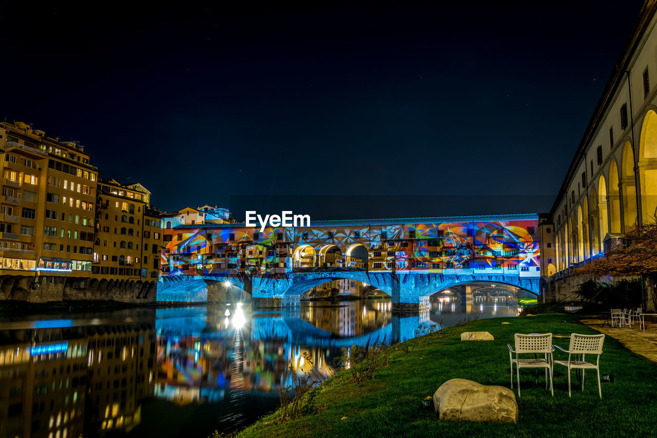 Illuminated bridge over river by buildings against clear sky at night