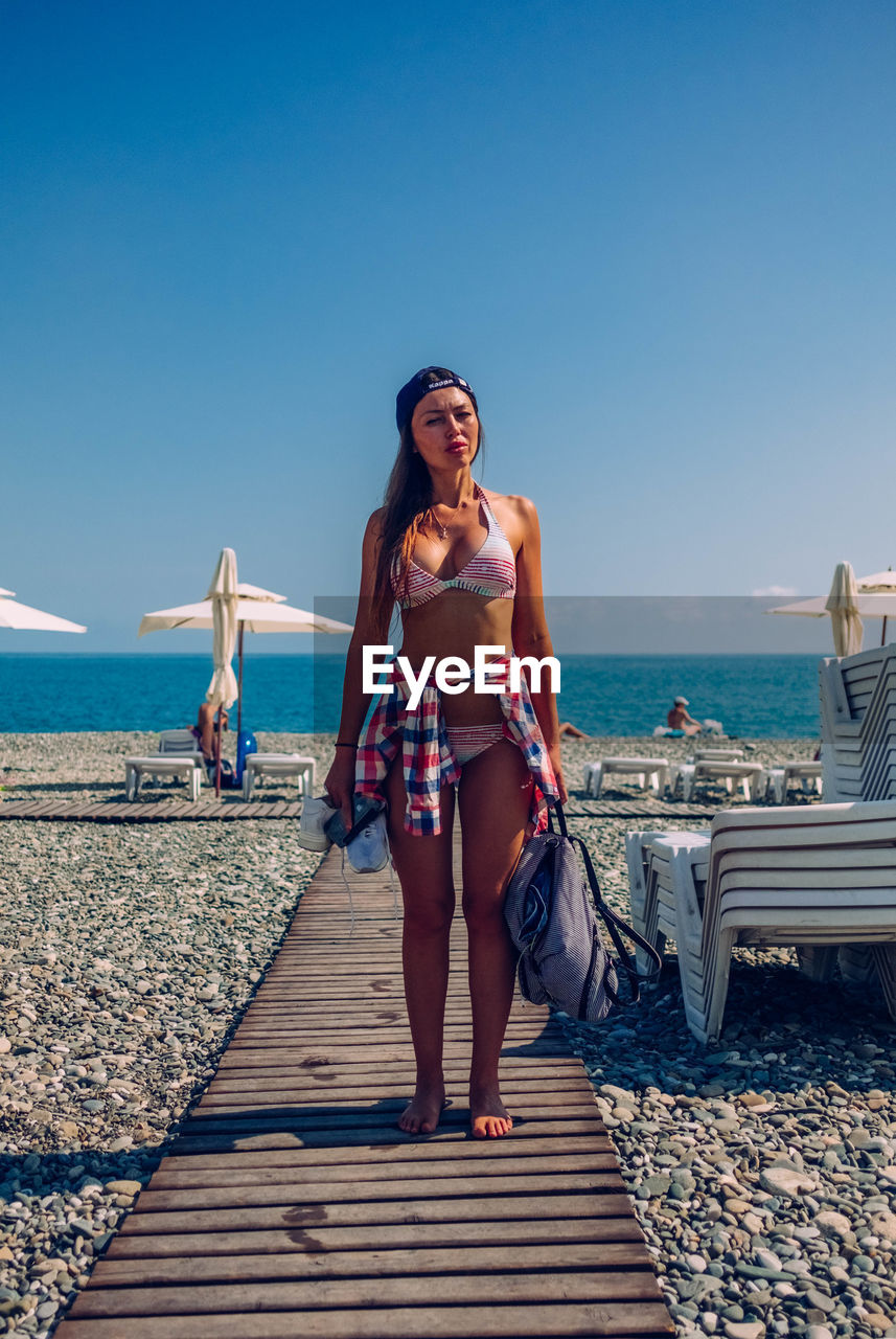 FULL LENGTH OF YOUNG WOMAN STANDING AT SEA SHORE AGAINST CLEAR SKY