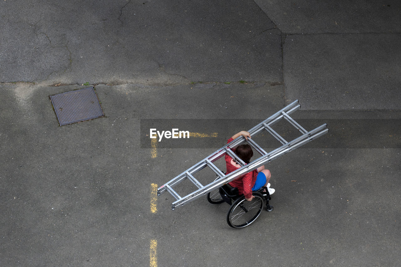 High angle view of girl on wheelchair carrying the ladder on street