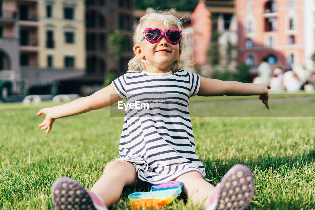 A funny little blonde in sunglasses and a striped dress sits on the lawn.