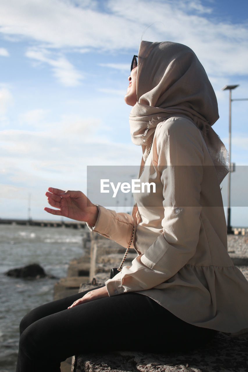 Side view of young woman sitting on retaining wall by sea against sky