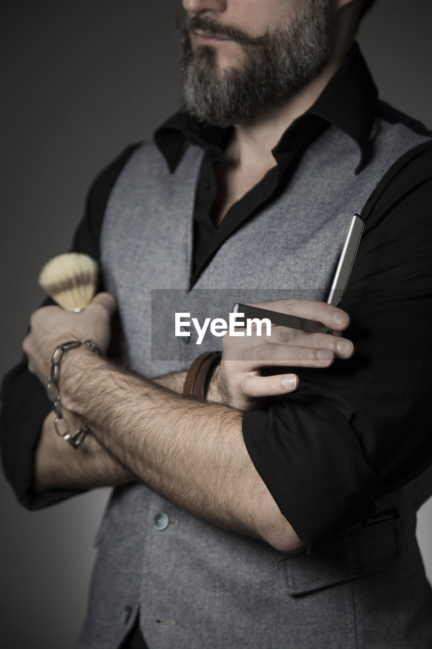 Midsection of man holding shaving brush and razor against gray background