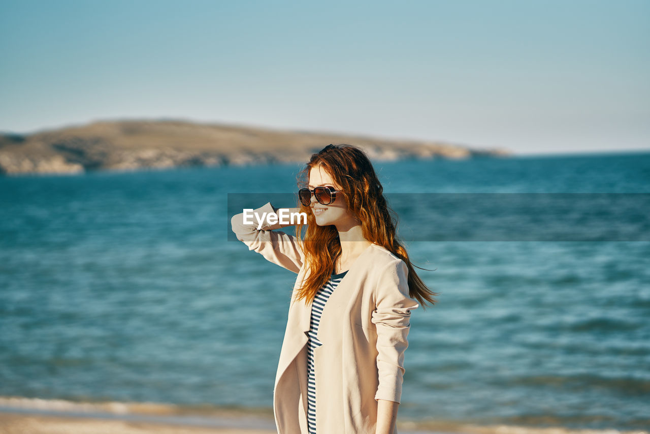 WOMAN WEARING SUNGLASSES STANDING AGAINST SEA