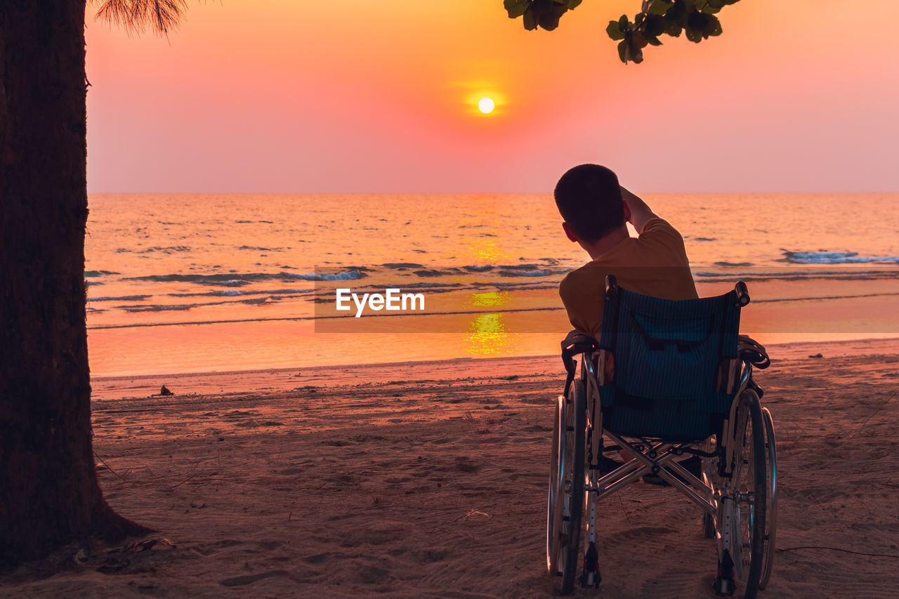 Rear view of man sitting in wheelchair on beach during sunset