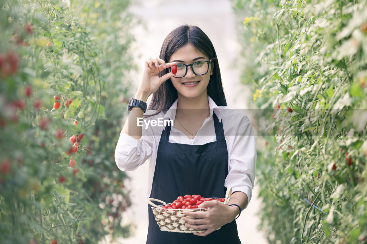 Portrait of smiling young woman holding fruits while standing amidst plants on field