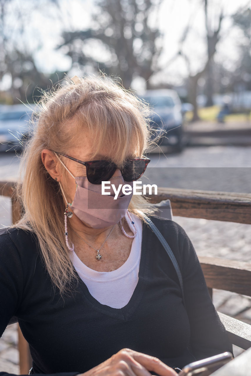 Mature adult blonde woman smiling with glasses wearing face mask texting to smartphone