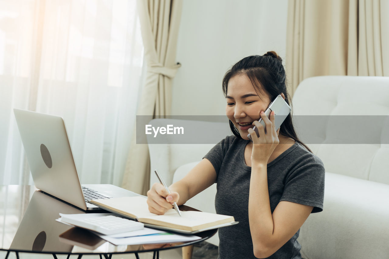 YOUNG WOMAN USING PHONE WHILE SITTING ON TABLE AT HOME