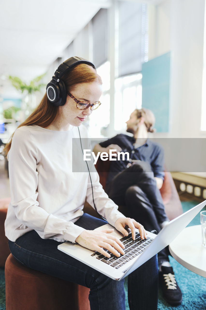 Businesswoman using laptop in creative office with colleague in background