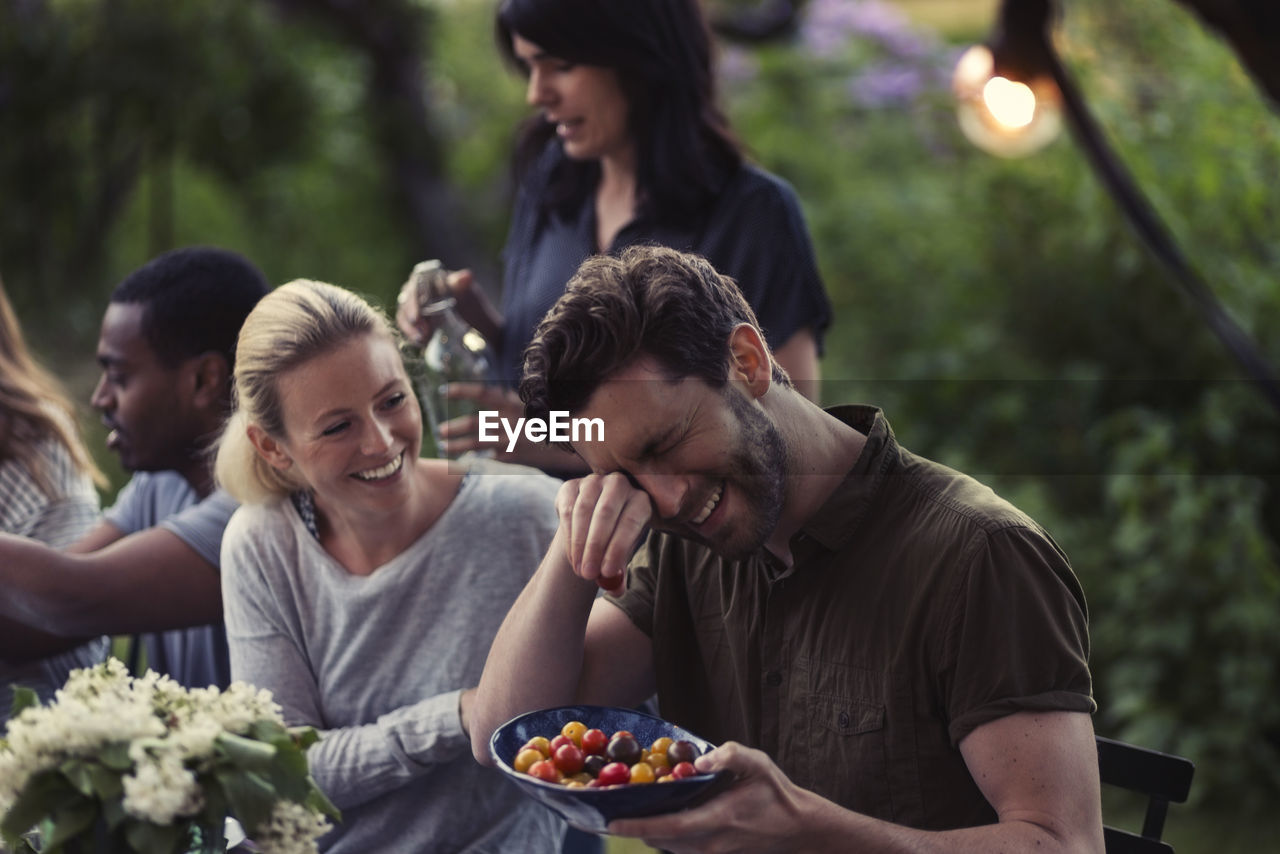 Happy woman looking at man rubbing eyes while holding tomato bowl at dinner party in yard