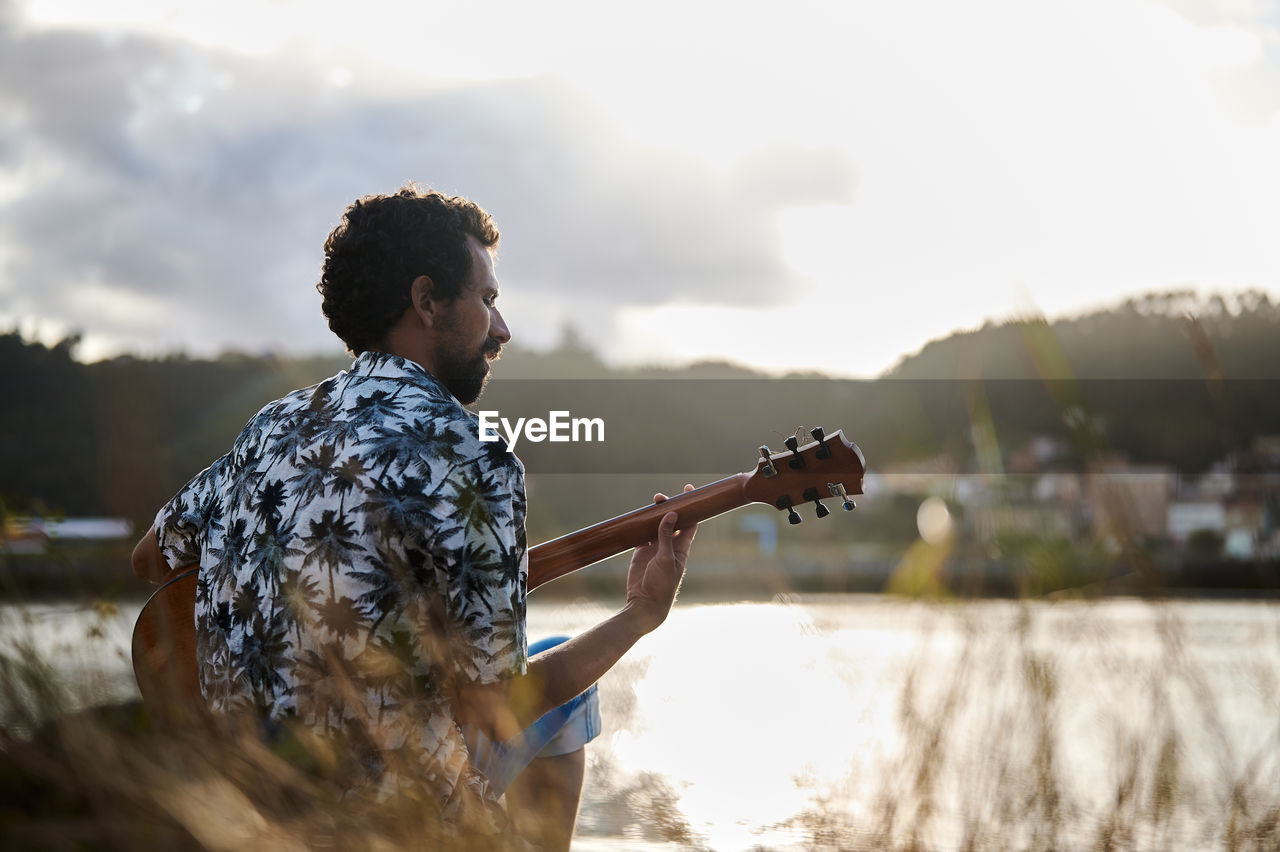 Back view of male musician playing acoustic guitar while sitting on seashore on sunny day