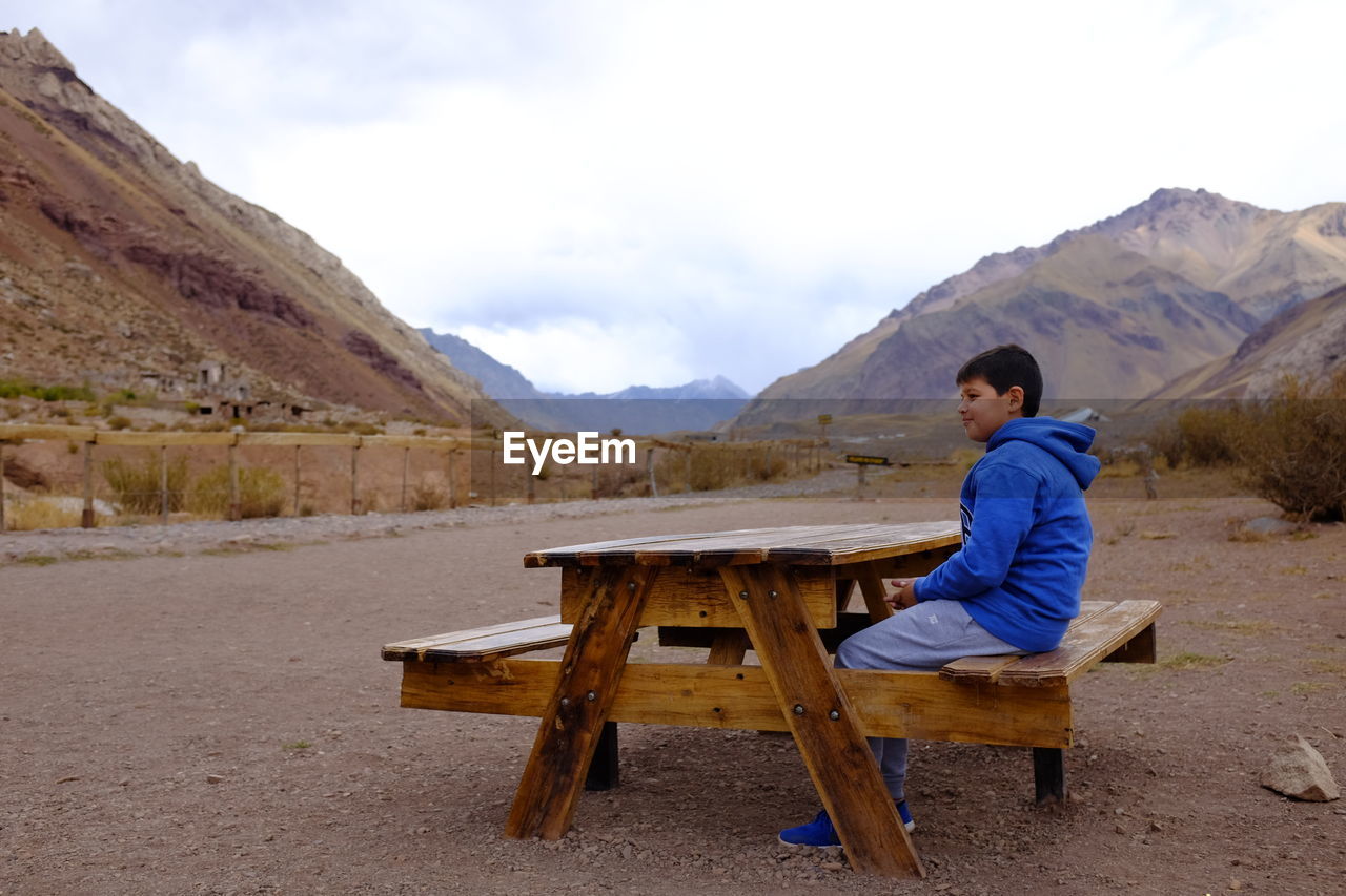 Boy looking away while sitting on seat against mountains