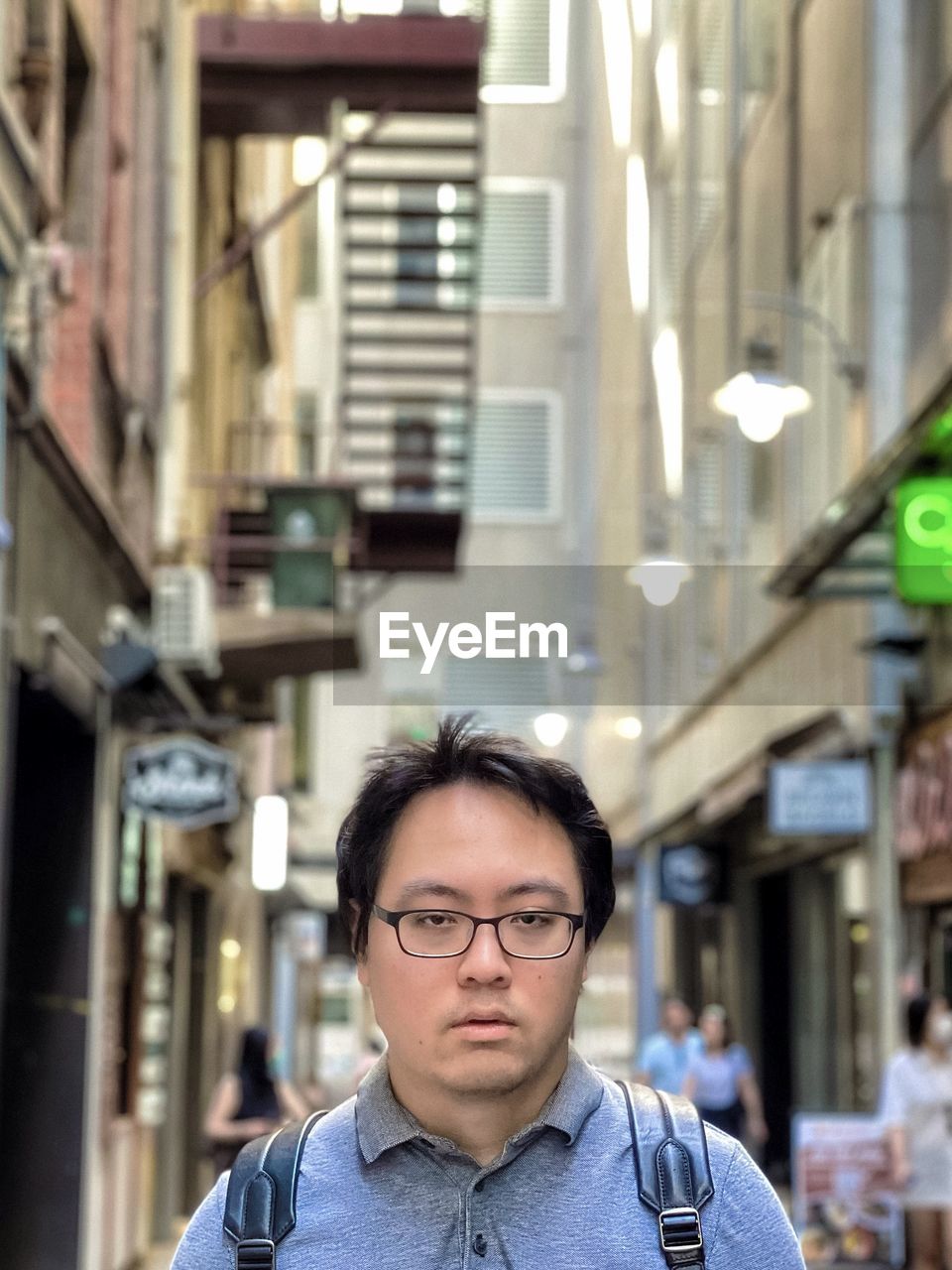 Portrait of young asian man in eyeglasses standing in laneway against buildings.