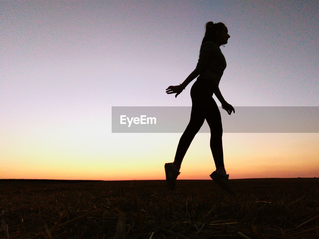 SILHOUETTE WOMAN WALKING ON FIELD AGAINST SKY DURING SUNSET