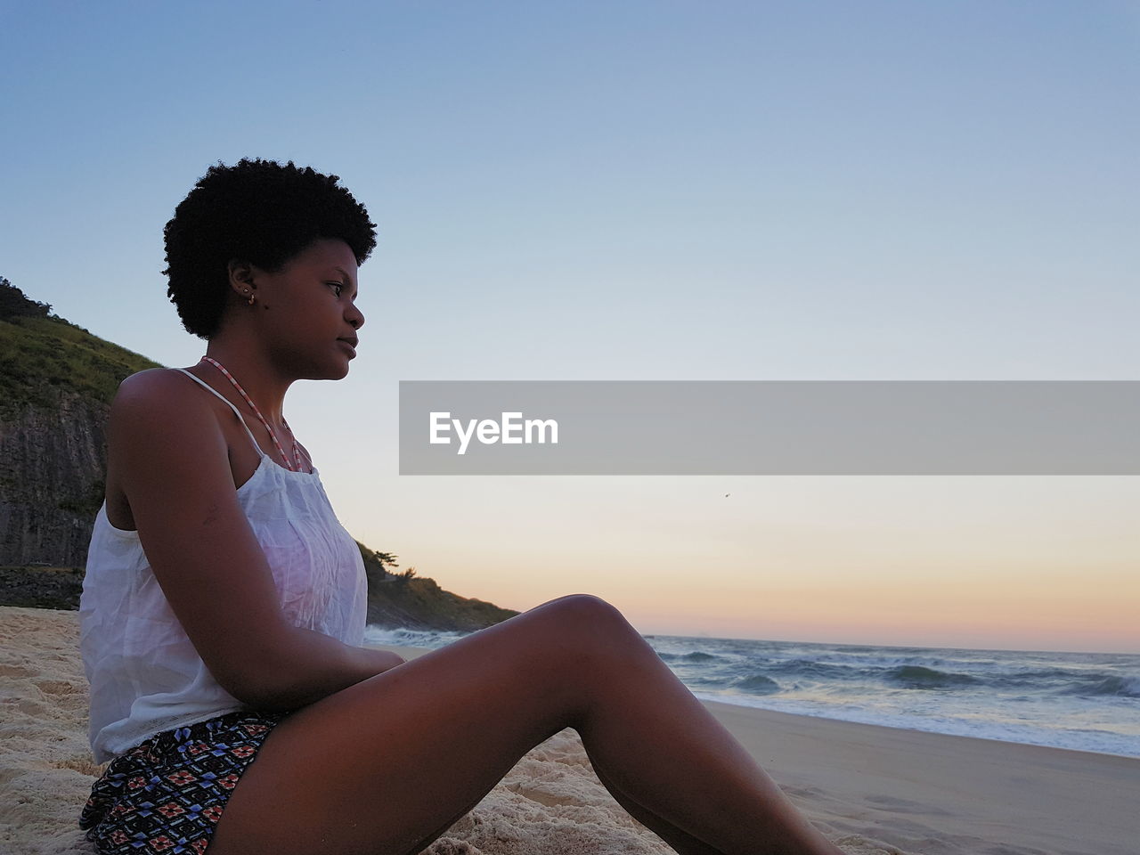 Woman looking away while sitting at beach against sky during sunset