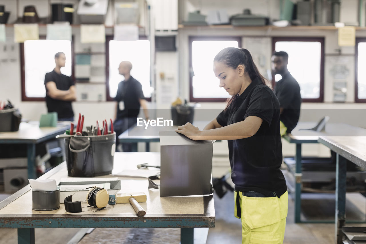 Side view of female student working on sheet metal with coworkers in background