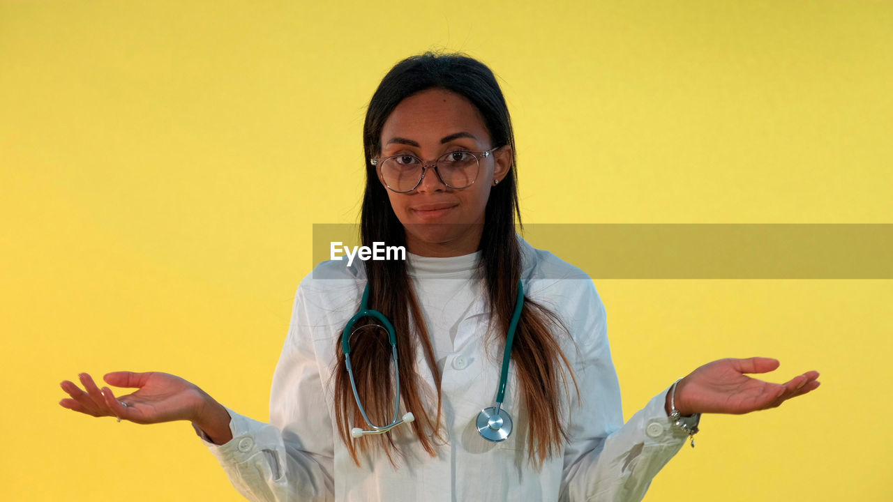 yellow, one person, colored background, yellow background, glasses, adult, eyeglasses, portrait, women, studio shot, waist up, long hair, hairstyle, indoors, looking at camera, person, front view, sign language, standing, female, smiling, young adult, copy space, clothing, healthcare and medicine, holding, black hair, emotion, gesturing, casual clothing