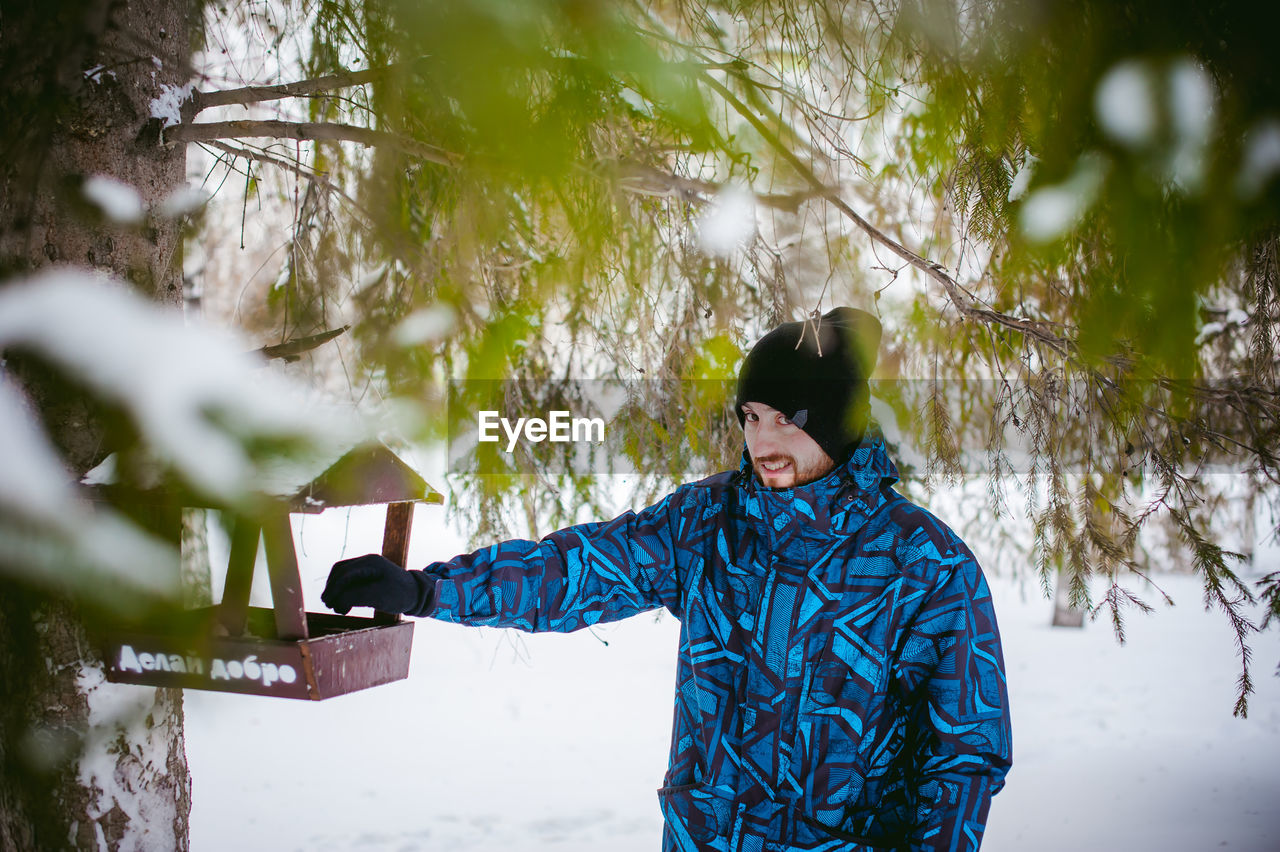 Portrait of young man standing by bird feeder on tree during winter