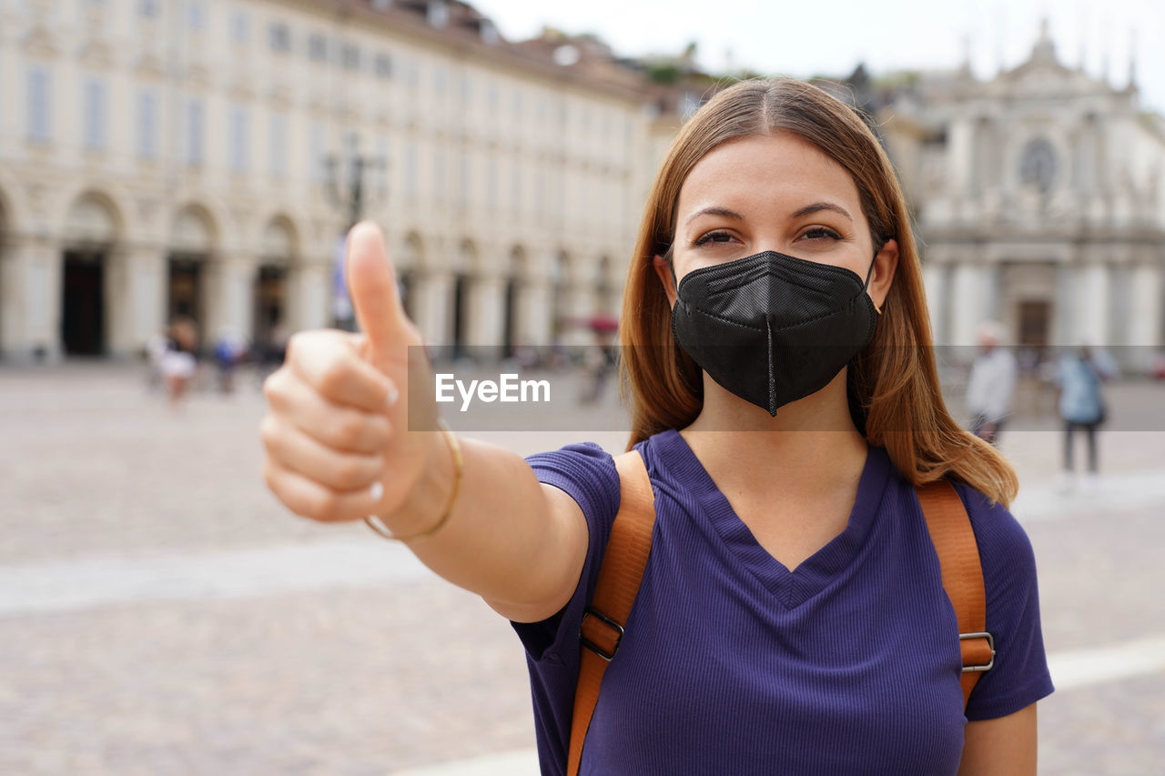 Optimistic traveler woman wearing black protective mask kn95 ffp2 showing thumbs up in city square