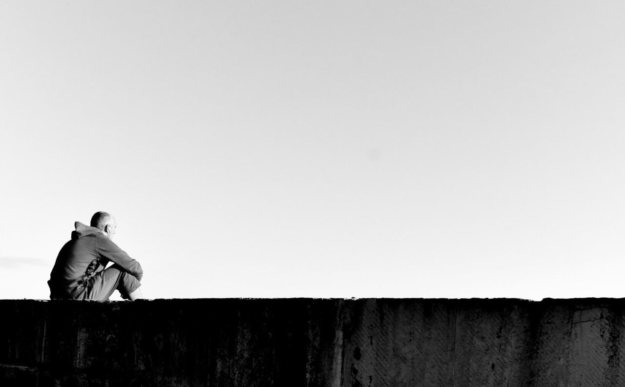 Man sitting on retaining wall against clear sky