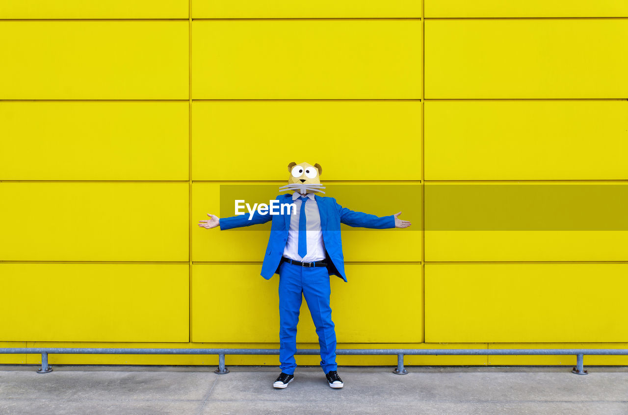 Man wearing vibrant blue suit and rodent mask standing in front of yellow wall with outstretched arms