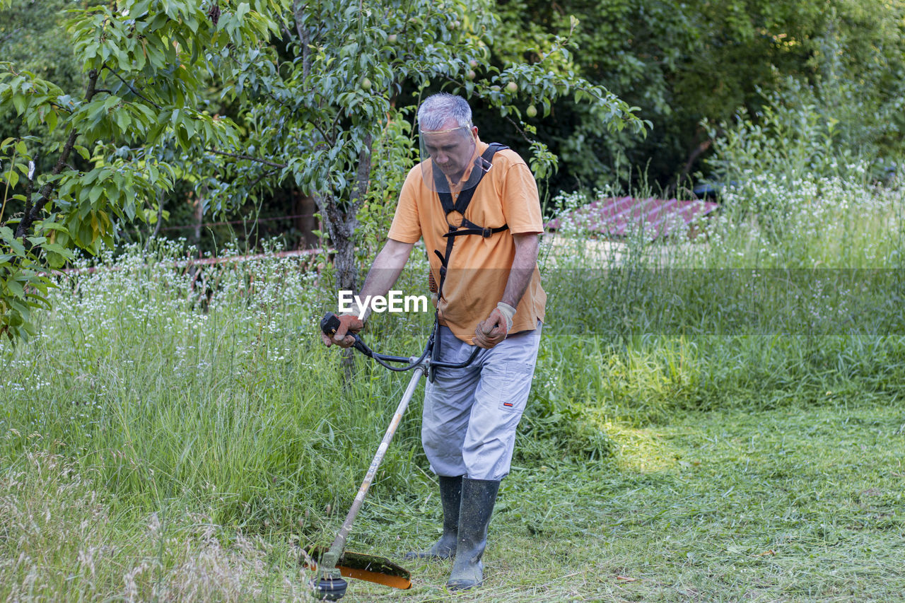 A mature man in protective clothing, and gloves  and mowing tall grass and the weeds with a trimmer