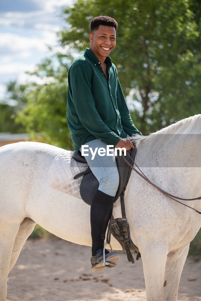 Smiling young man riding horse against tree