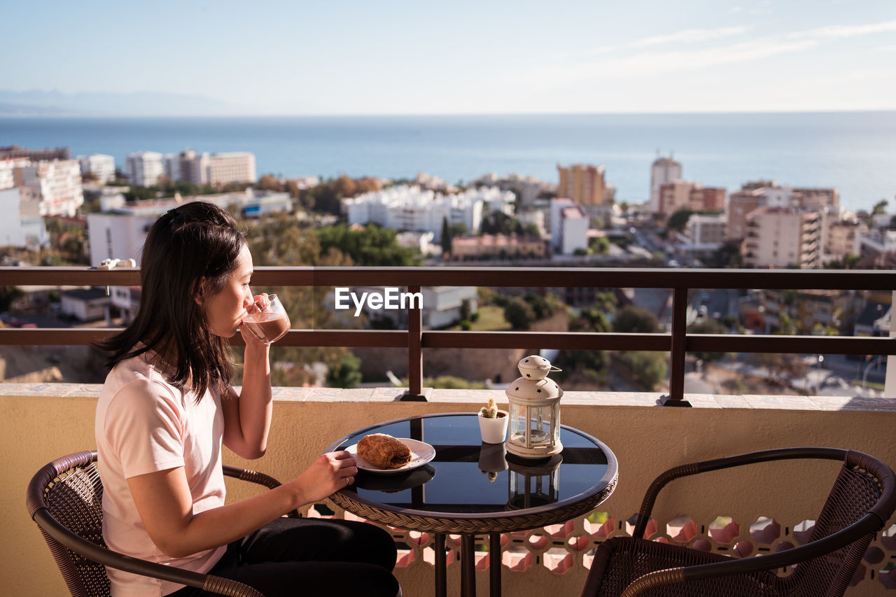 Happy asian female with glass of hot drink in hand looking at camera while sitting on balcony at table with food during breakfast