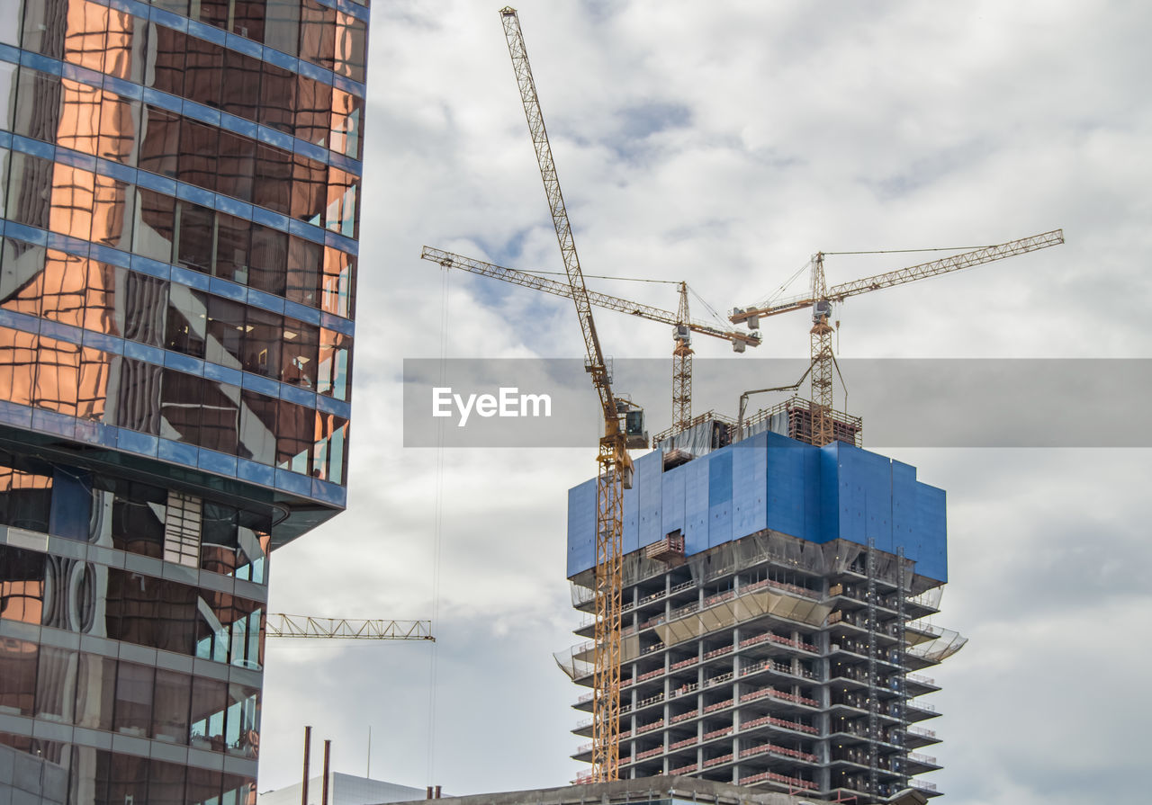 Construction of a skyscraper with three tower cranes against a cloudy sky.