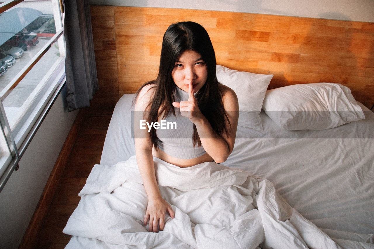 Portrait of young woman with finger on lips sitting on bed