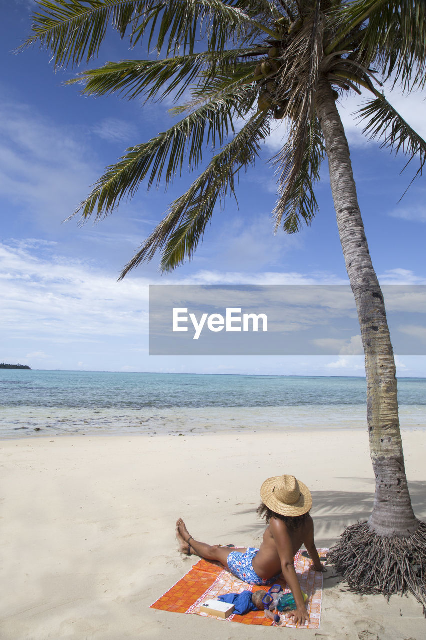 Man with sunhat and curly hair chilling under palm tree at sandy beach