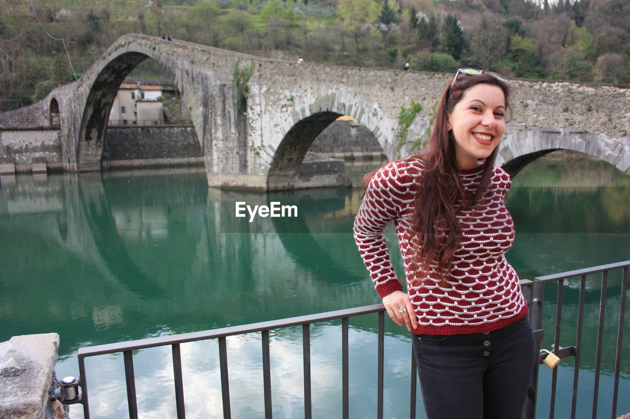 SMILING YOUNG WOMAN STANDING ON FOOTBRIDGE AGAINST WATER