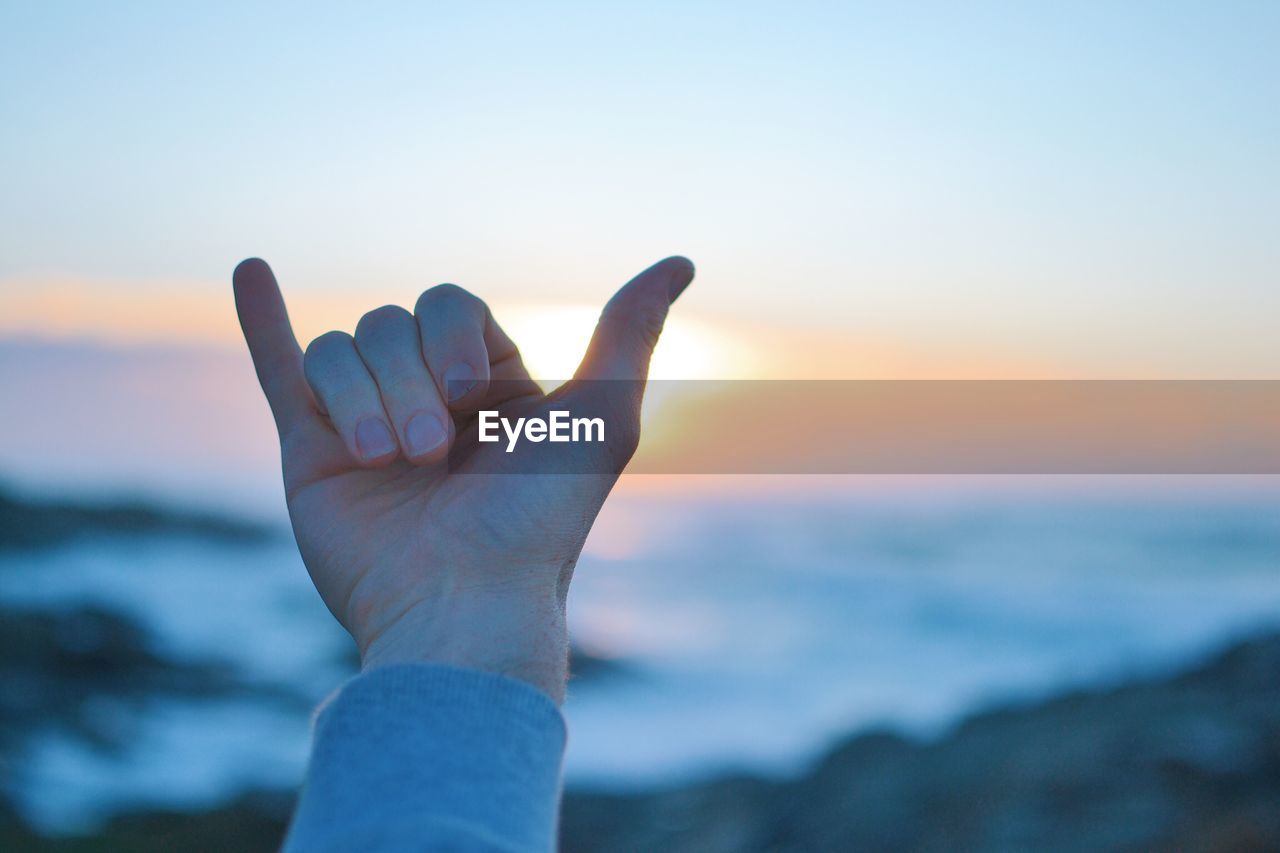 Cropped hand of person gesturing horn sign at beach during sunset