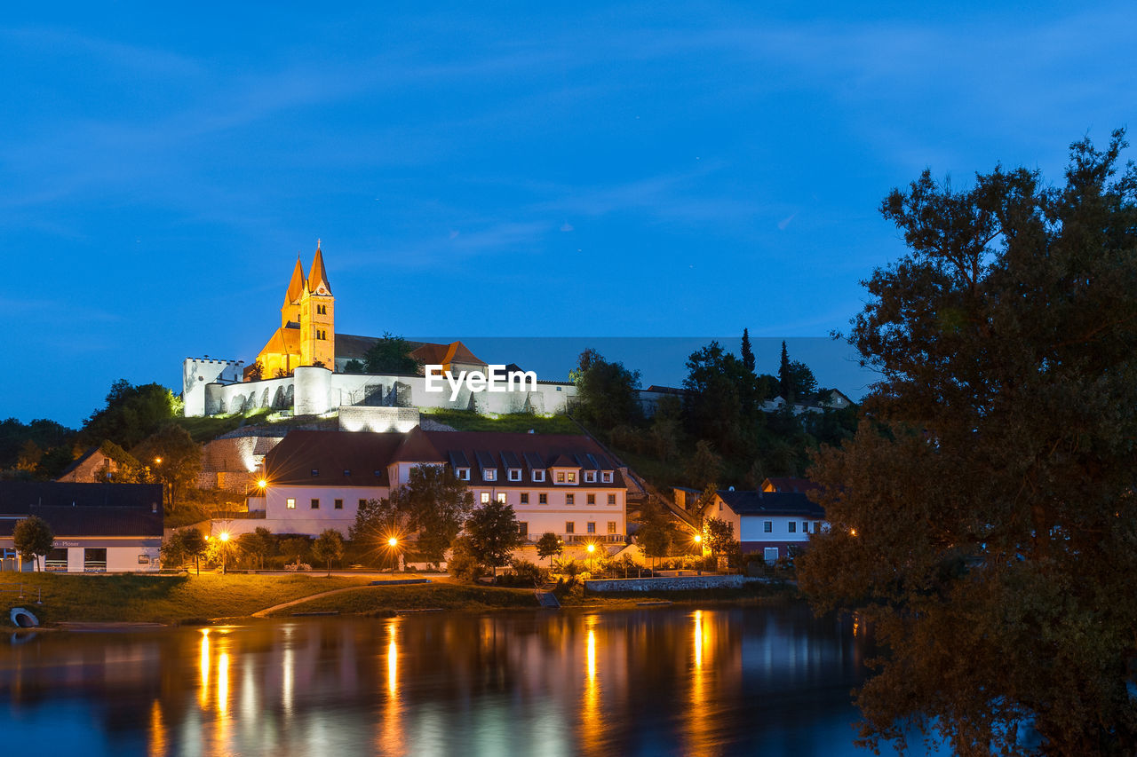 Illuminated buildings by river against sky at dusk