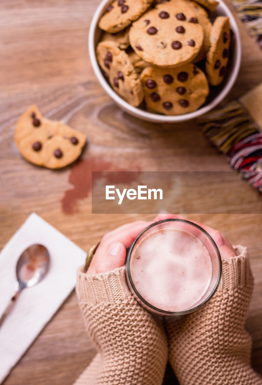 Closeup of female hands holding hot drink glass over table with chocolate chip cookies on stars bowl